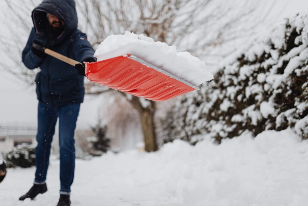 A person using a red shovel for snow removal on a snowy day, with snow-covered trees in the background.