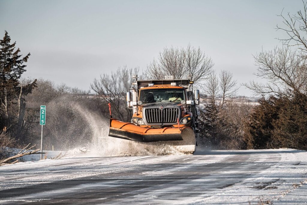 An orange snowplow engaged in snow clearing on a snowy road amidst a winter landscape, with trees on either side and a blue sky above.